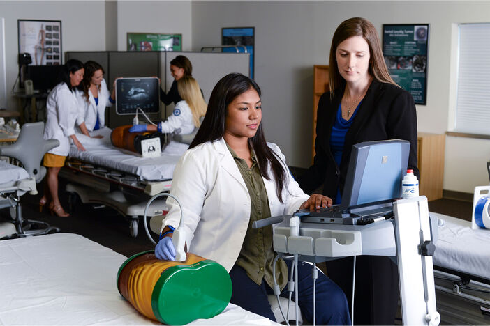 A female sonography student practices scanning an abdomen phantom while a faculty supervises